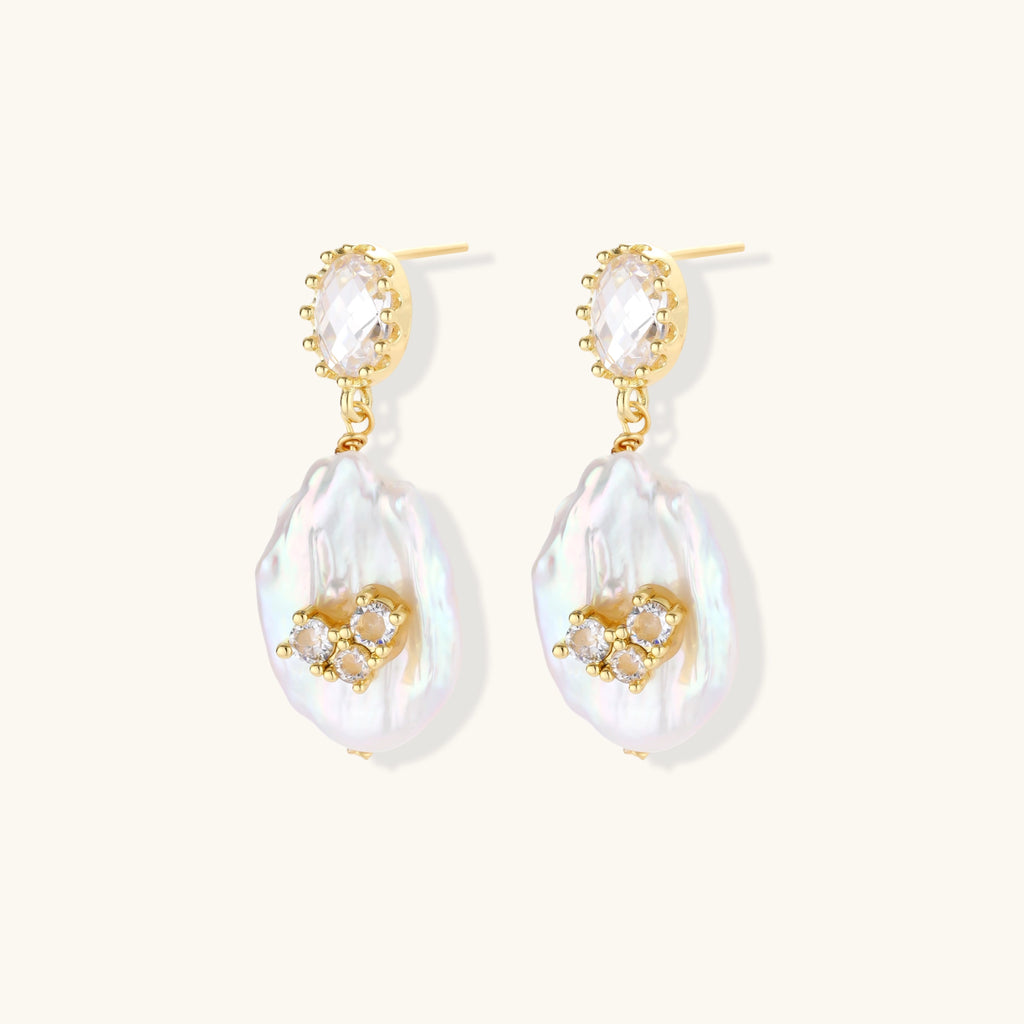 Gold-plated drop earrings with iridescent baroque pearls and crystal flower accents on cream background