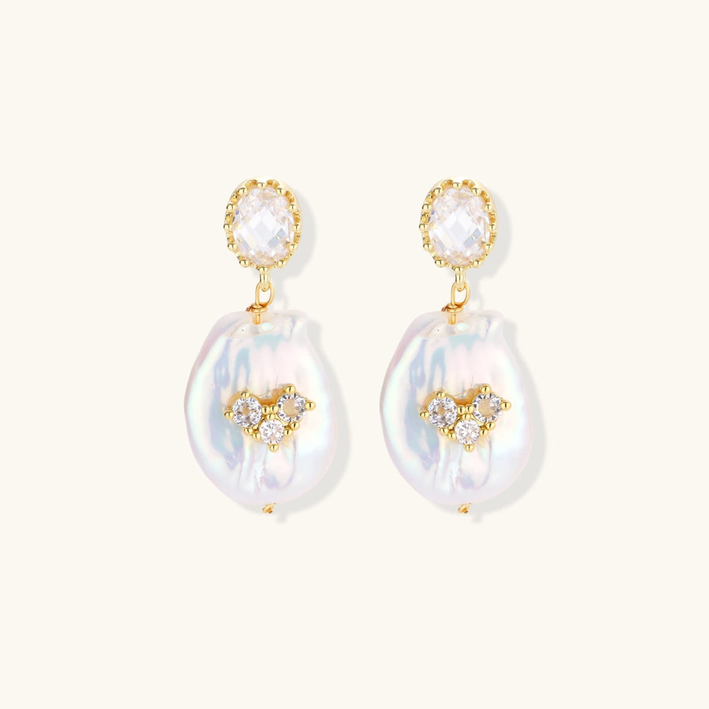 Gold-plated drop earrings with iridescent baroque pearls and crystal flower accents on cream background
