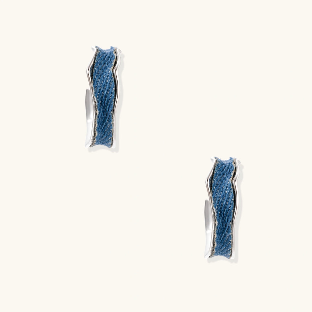 Silver huggie hoops with blue denim fabric inlay strip and polished white gold plated finish on cream background.
