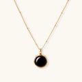 Circular black agate pendant on gold-plated sterling silver chain against cream background, showing modern minimalist design.