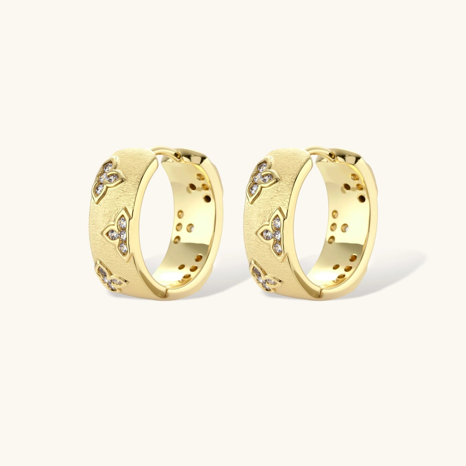 Gold-plated hoop earrings with textured surface and raised floral motifs set with clear zirconia stones on cream background.