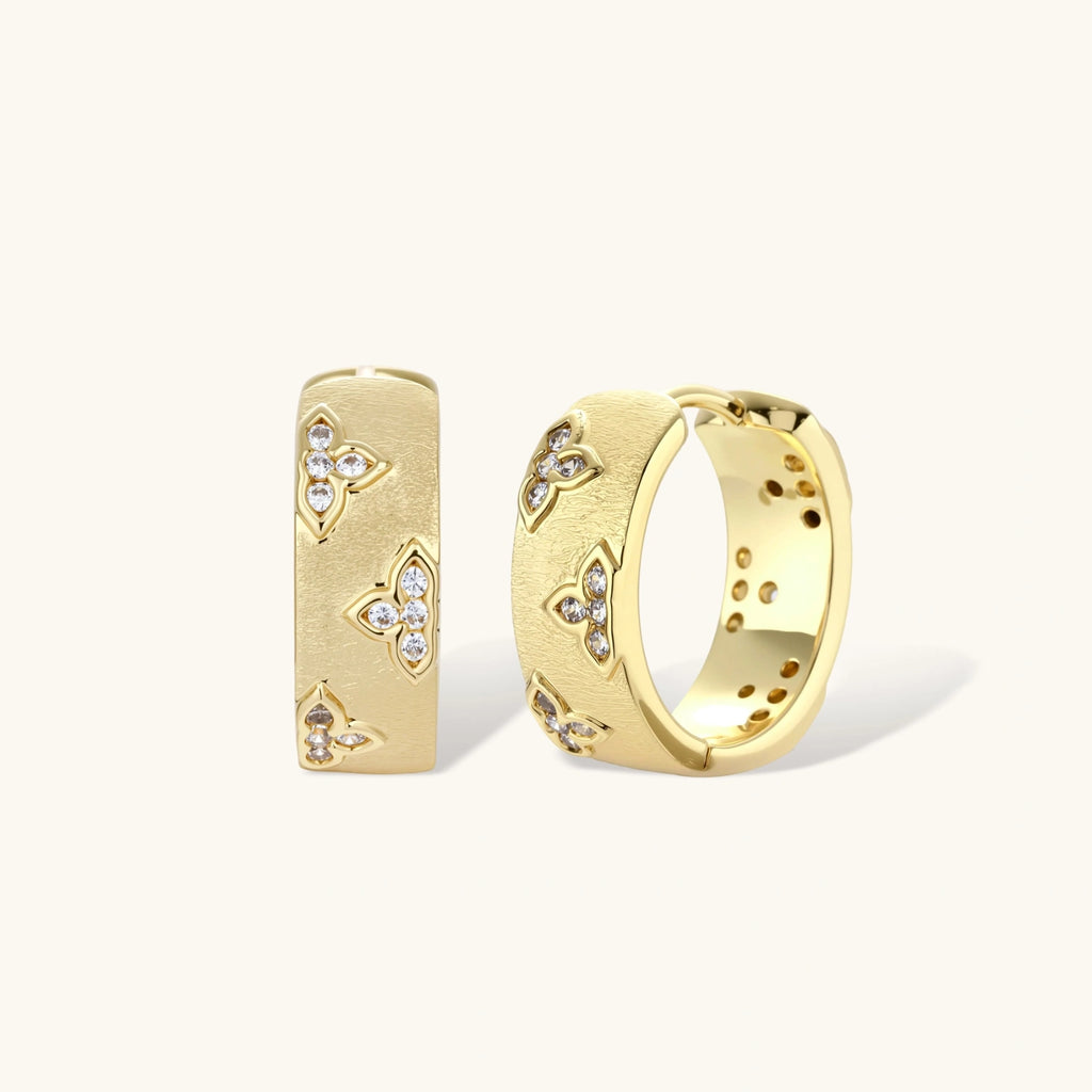 Gold-plated hoop earrings with textured surface and raised floral motifs set with clear zirconia stones on cream background.