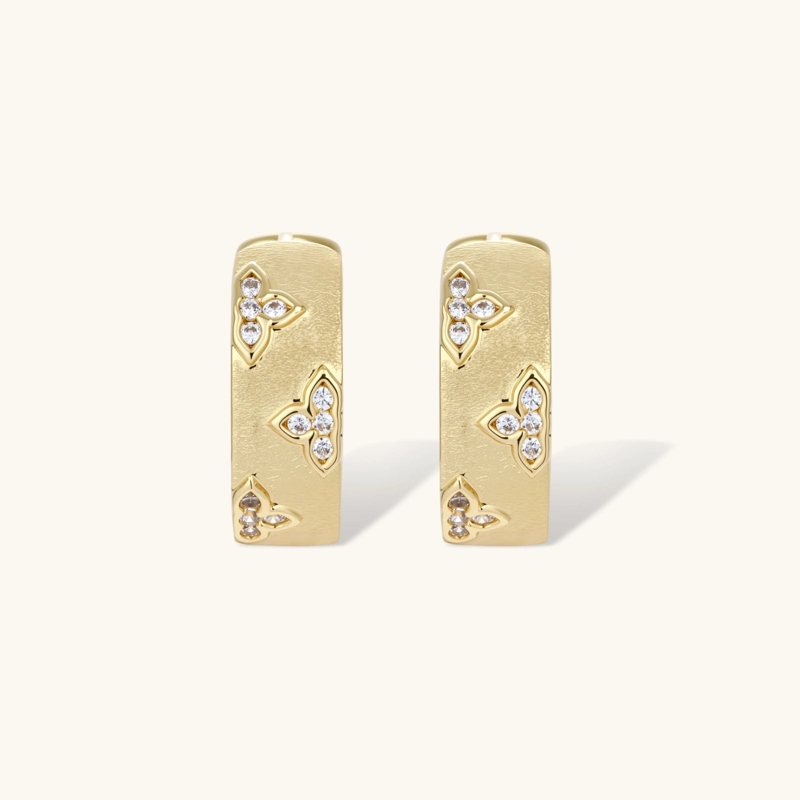 Gold-plated hoop earrings with textured surface and raised floral motifs set with clear zirconia stones on cream background.