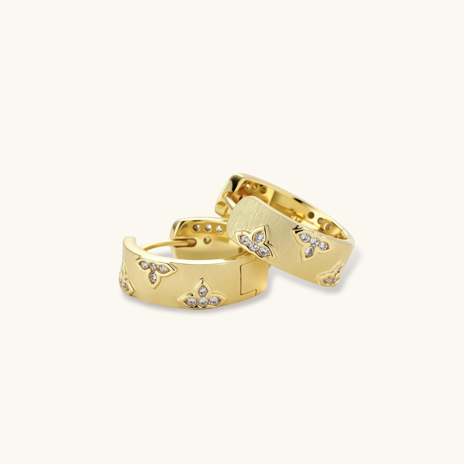 Gold-plated hoop earrings with textured surface and raised floral motifs set with clear zirconia stones on cream background.