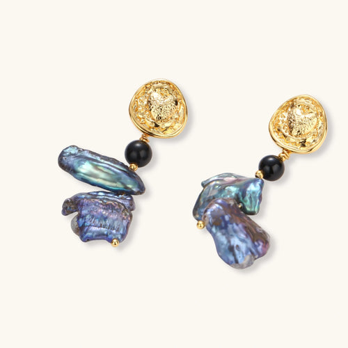 Ornate gold-plated drop earrings with black beads and iridescent blue-purple baroque pearls against white background.
