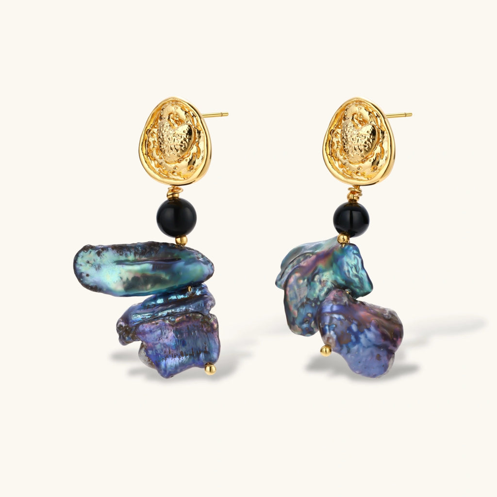 Ornate gold-plated drop earrings with black beads and iridescent blue-purple baroque pearls against white background.